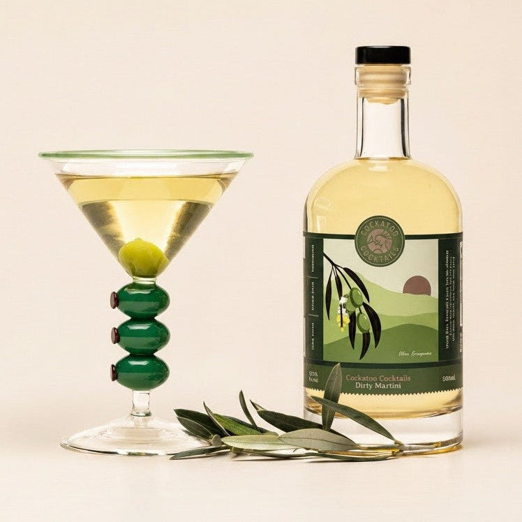 A bottle of Dirty Martini stands next to a dirty martini glass filled with a light yellow cocktail and a green olive on a stem, with olive leaves in front, on a warm beige background.