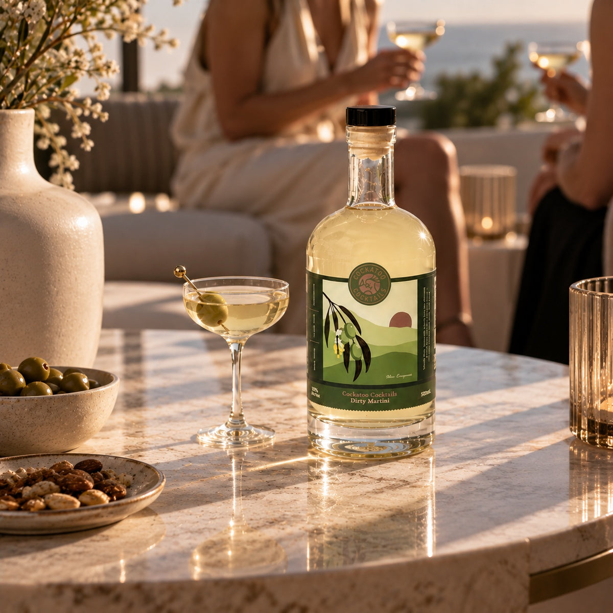 A bottle of Dirty Martini sits on a marble table in a sunlit outdoor setting, surrounded by a cocktail, olives, nuts, and blurred people in the background.
