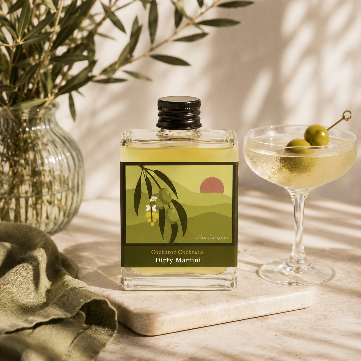 A clear glass bottle of dirty martini cocktail with a black cap and a green-and-yellow illustrated label reading 'Dirty Martini,' styled on a marble board beside a coupe glass with olives, with olive branches and draped fabric in the background.