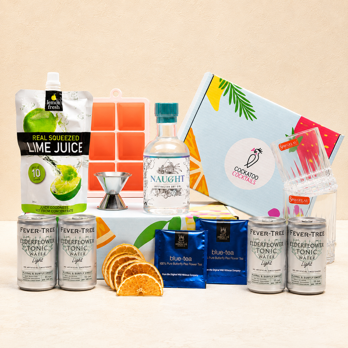 A cocktail gift set arranged on a light beige surface against a warm beige background. The set includes a pouch of lime juice, a bottle of Naught Gin, multiple cans of Fever Tree tonic, butterfly pea tea packets, orange slices, a metal jigger, a colorful ice cube tray, a Cockatoo Cocktails branded gift box, a jigger and two glasses. 