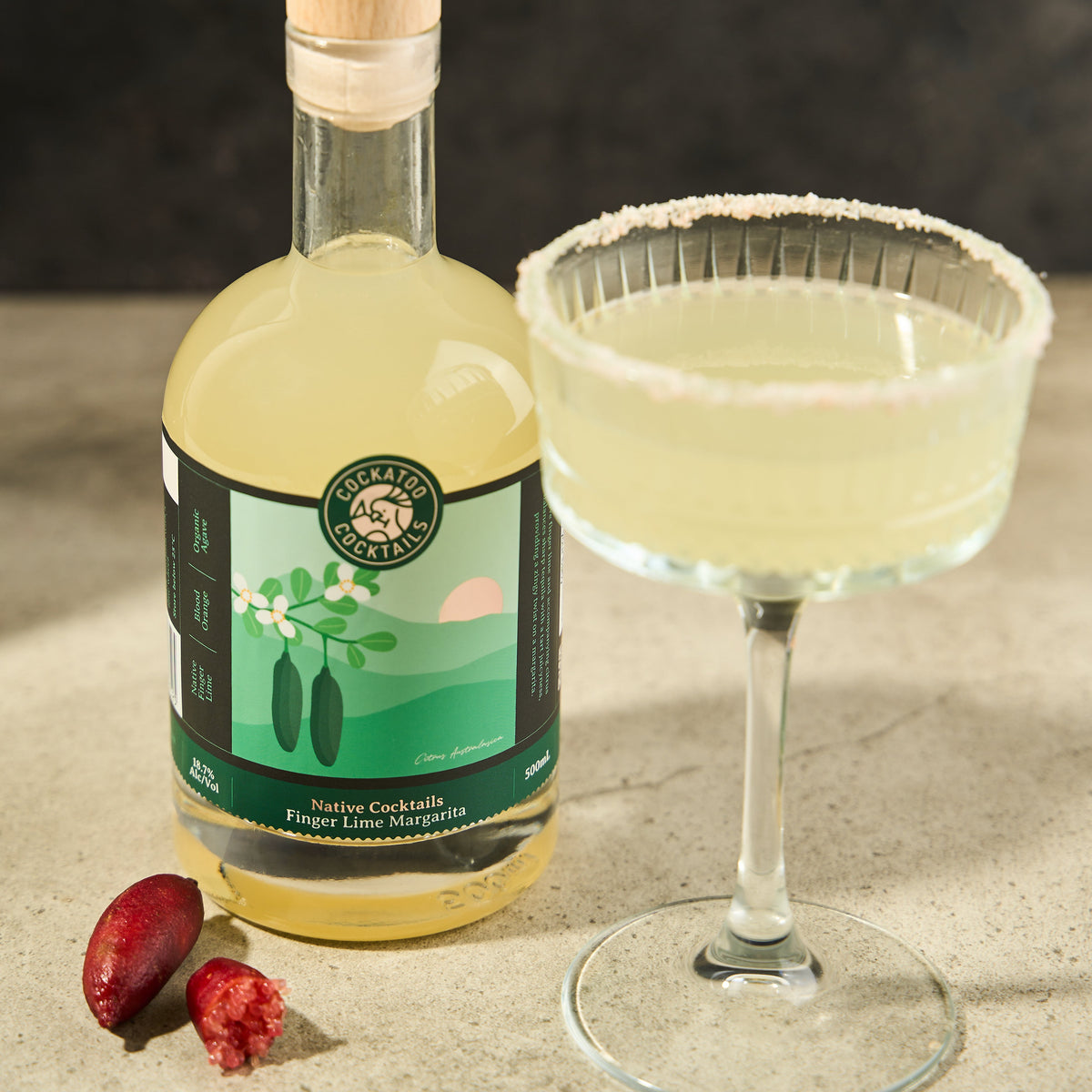 A bottle of Finger Lime Margarita Cocktail with a green label and a clear glass coupe, set on a stone surface with a dark textured background. Two finger limes sit beside the bottle.