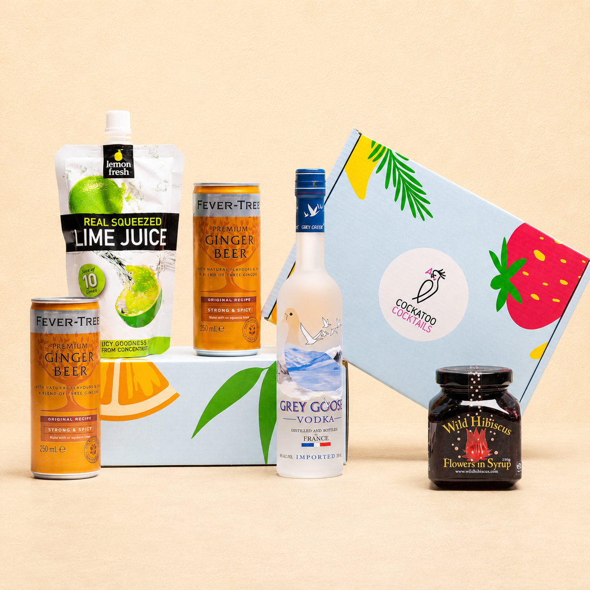 A curated gift set featuring Fever-Tree ginger beer, lime juice, Grey Goose vodka, a jar of Wild hibiscus flowers and a cockatoo cocktails branded gift box, arranged on a light beige background.