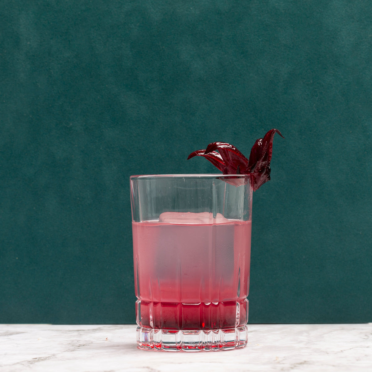 A clear glass tumbler filled with a pinkish-red drink and a large ice cube, garnished with a dark red hibiscus flower, set on a white marble surface against a dark teal-green background.