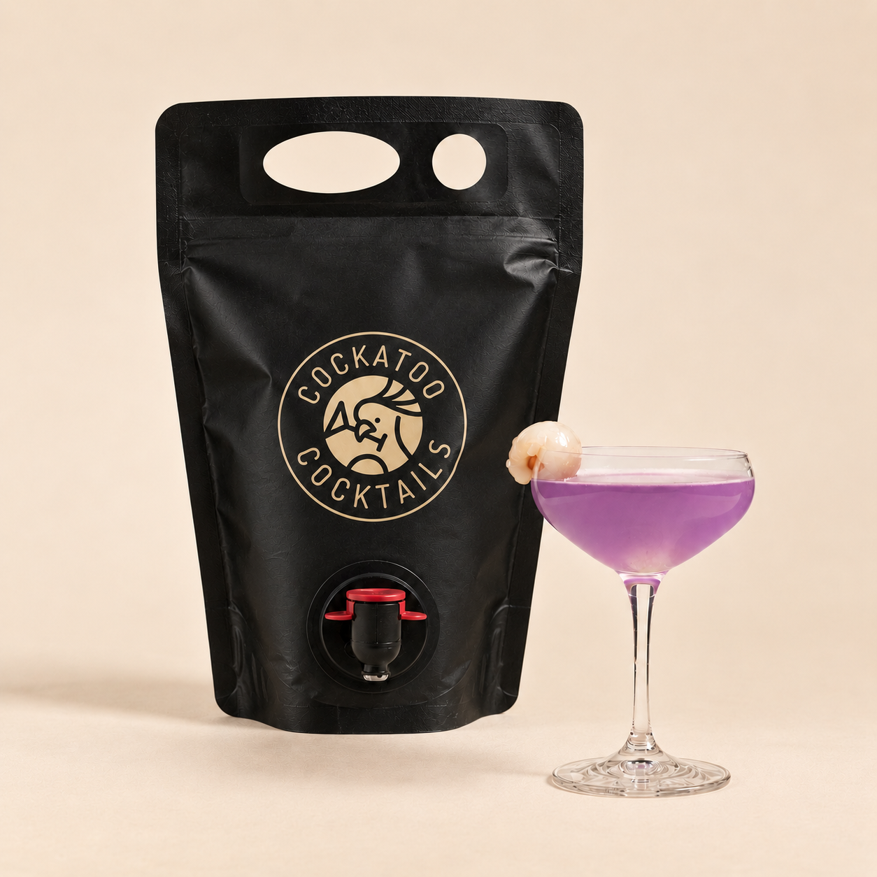 A black stand-up pouch with a handle and a small spout, branded 'Cockatoo Cocktails' in beige, shown next to a coupe glass filled with a purple lychee martini cocktail on a warm beige background.