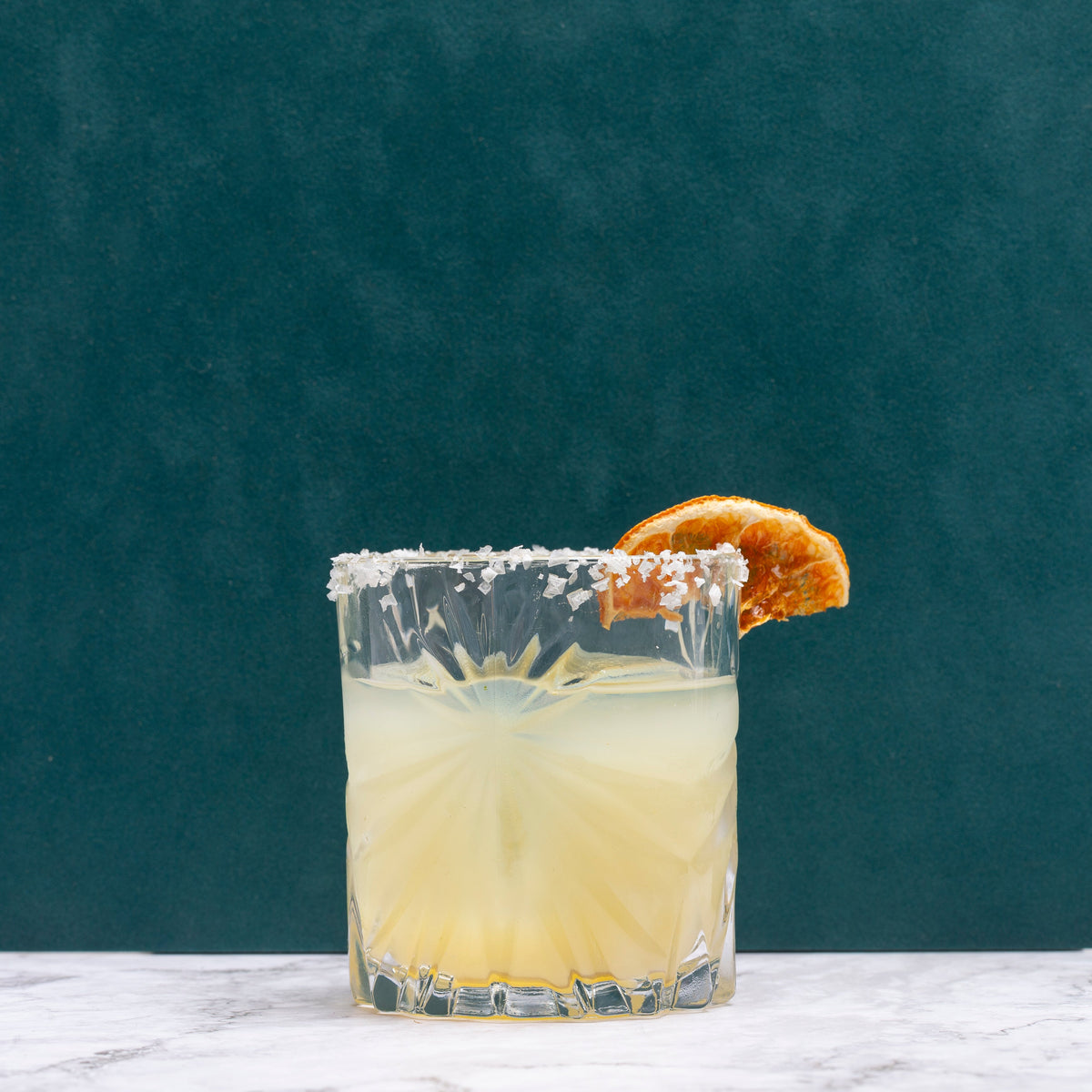 A clear cut-glass tumbler filled with a Margarita, rimmed with coarse white salt, and garnished with a dried orange slice, set on a light marble surface against a dark teal background.