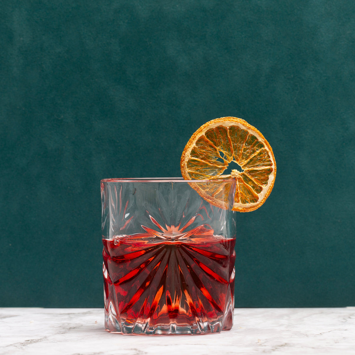 A clear cut-glass tumbler filled with a Negroni, set on a white marble surface against a dark teal background, with a dried orange slice garnish resting on the rim.