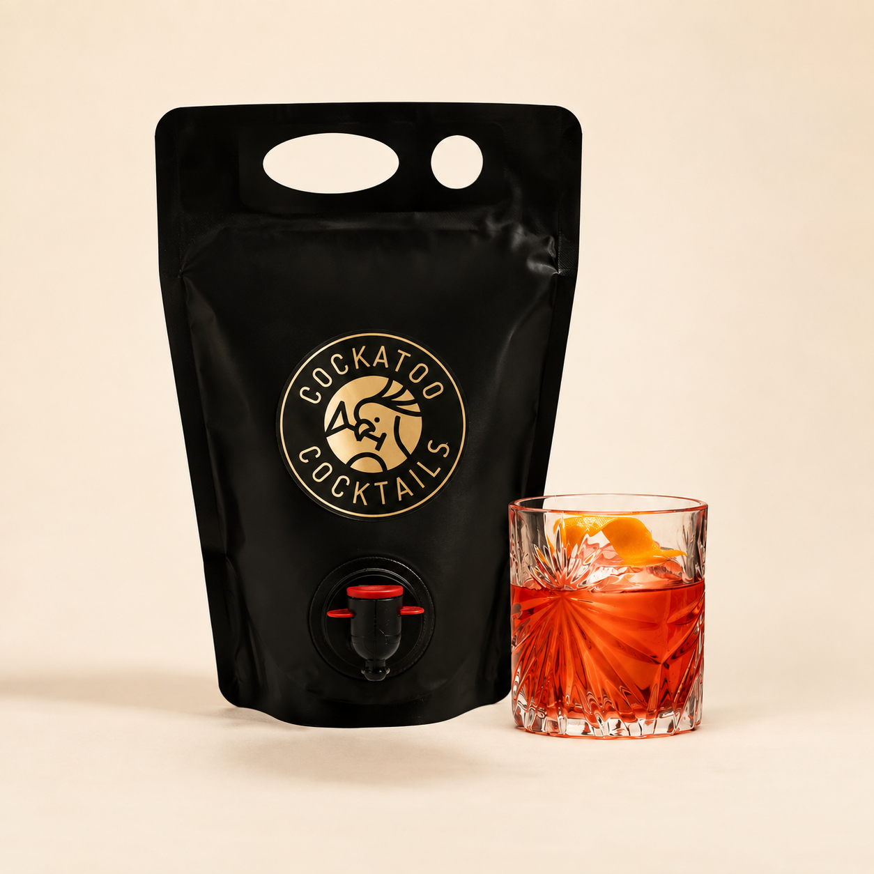 A black stand-up pouch with Negroni with a gold circular Cockatoo Cocktails logo and a small dispensing spout, shown next to a cut-glass tumbler filled with Negroni cocktail on a warm beige background.