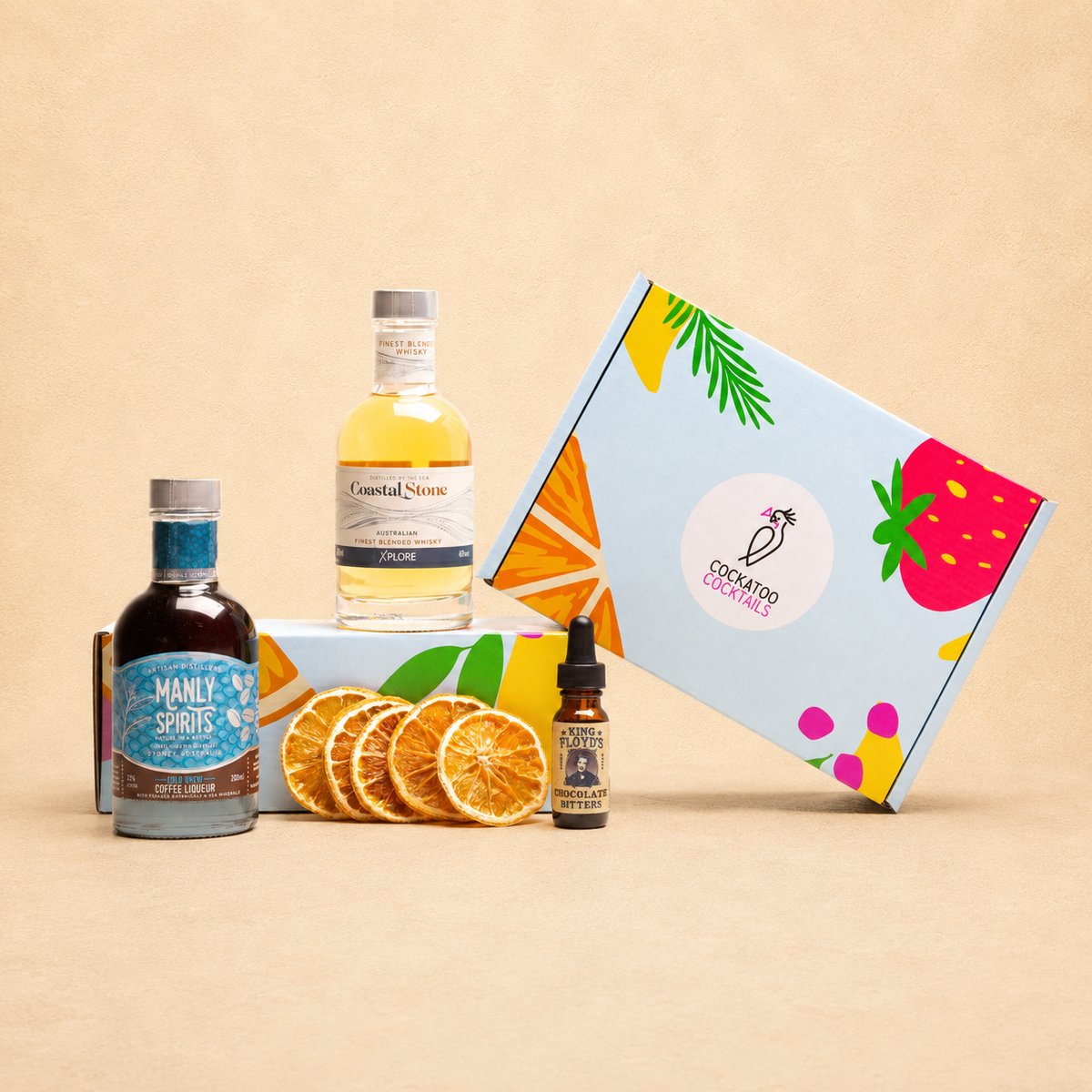 A curated gift set featuring a bottle of Manly's coffee liqueur, a bottle of Coastal Stone whisky, bitters, and orange slices arranged in front of a colorful Cockatoo Cocktails gift box on a beige background.