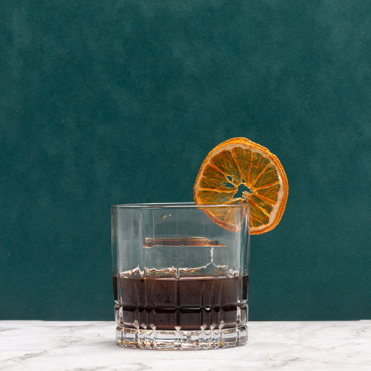 A clear cut-glass tumbler filled with an old fashioned cocktail, garnished with a dried orange slice, set on a white marble surface against a dark teal-green textured background.