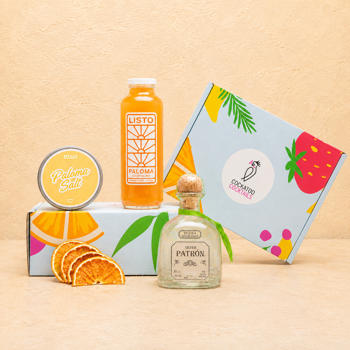 A curated gift set featuring a bottle of Listo Paloma, a bottle of Patrón tequila,  Paloma Salt, and colorful branded Cockatoo Cocktails gift box on a beige textured background.