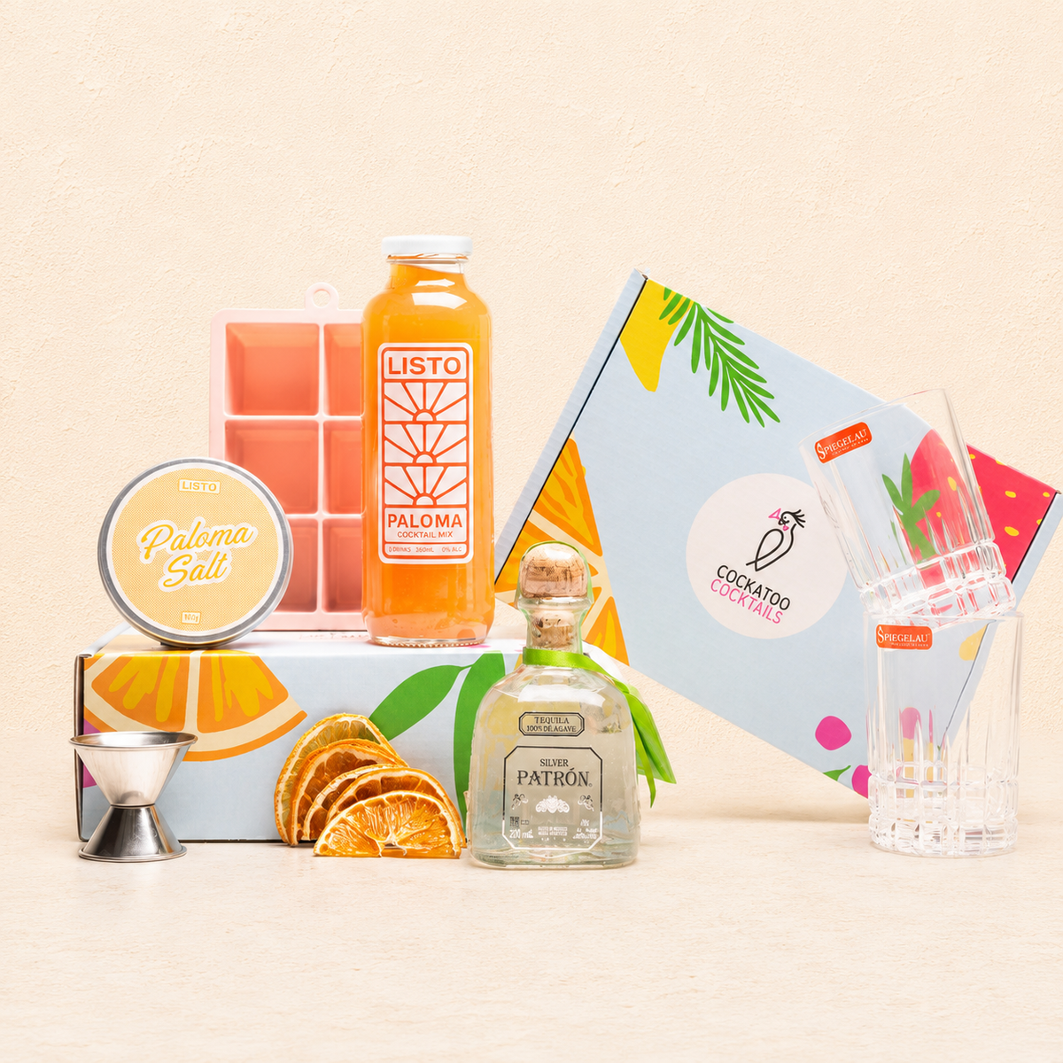 A curated cocktail gift set arranged on a light beige surface against a matching beige background. The set includes a bottle of Paloma Mixer, a bottle of Patrón tequila, a tin of Paloma Salt, a jigger, dehydrated orange slices, a colorful Cockatoo Cocktails gift box, and two  glasses with a jigger. 