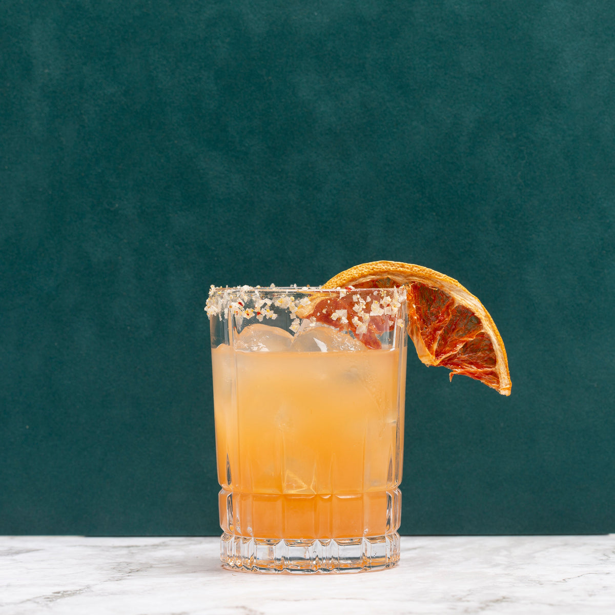 A Paloma rimmed with coarse salt and garnished with a dried orange slice, set on a white marble surface against a dark teal background.