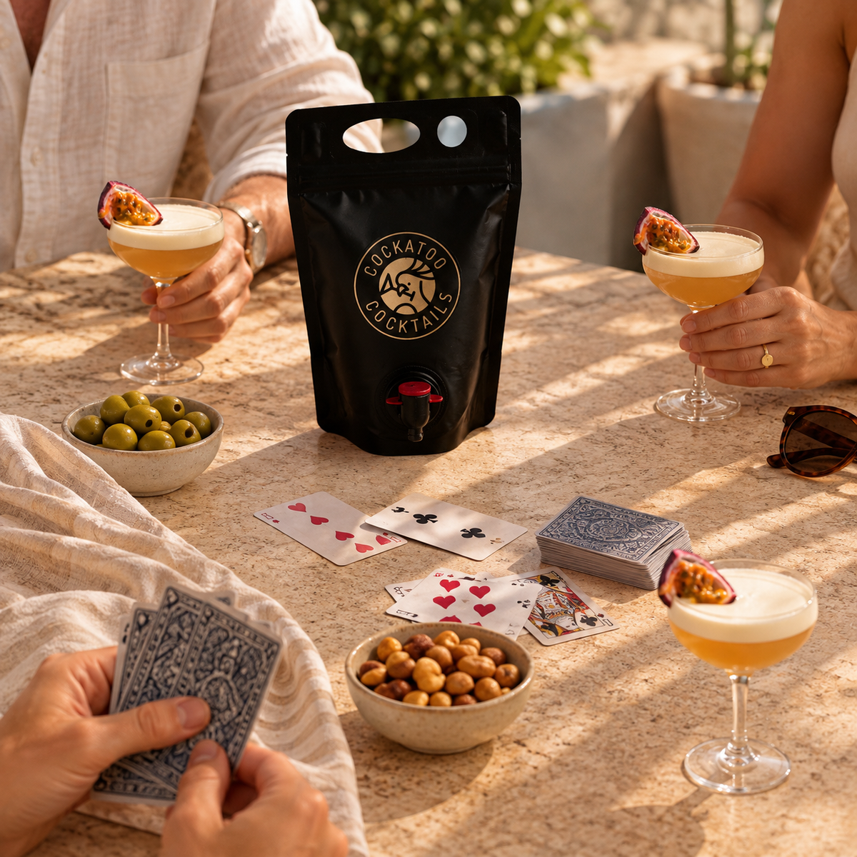 A black stand-up pouch of Cockatoo Cocktails is centered on a sunlit table, surrounded by people holding passionfruit martini glasses, bowls of olives and chickpeas, playing cards, and a lemon garden in the background.
