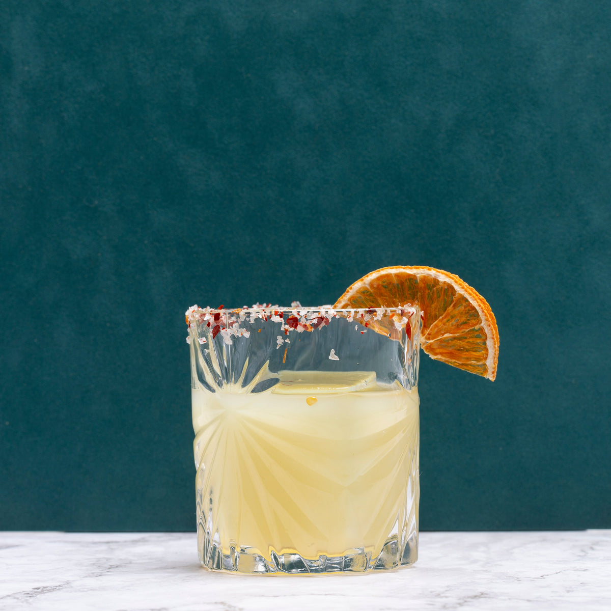 A clear cut-glass tumbler filled with a spicy margarita drink, rimmed with salt and garnished with a dried orange slice, set on a light marble surface against a dark teal background.