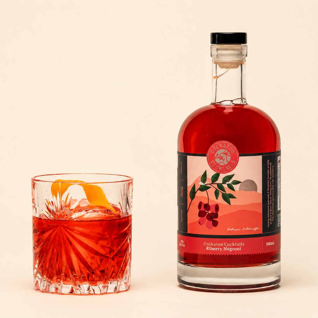 A bottle of Riberry Negroni next to a cut-glass tumbler filled with the same red cocktail and garnished with an orange peel, on a warm cream background.