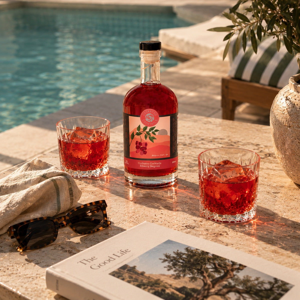A bottle of Riberry Negroni with a black cap and a pink label sits on a stone patio beside a swimming pool, with two rocks glasses filled with the red liquid, sunglasses, a folded towel, and a book nearby.