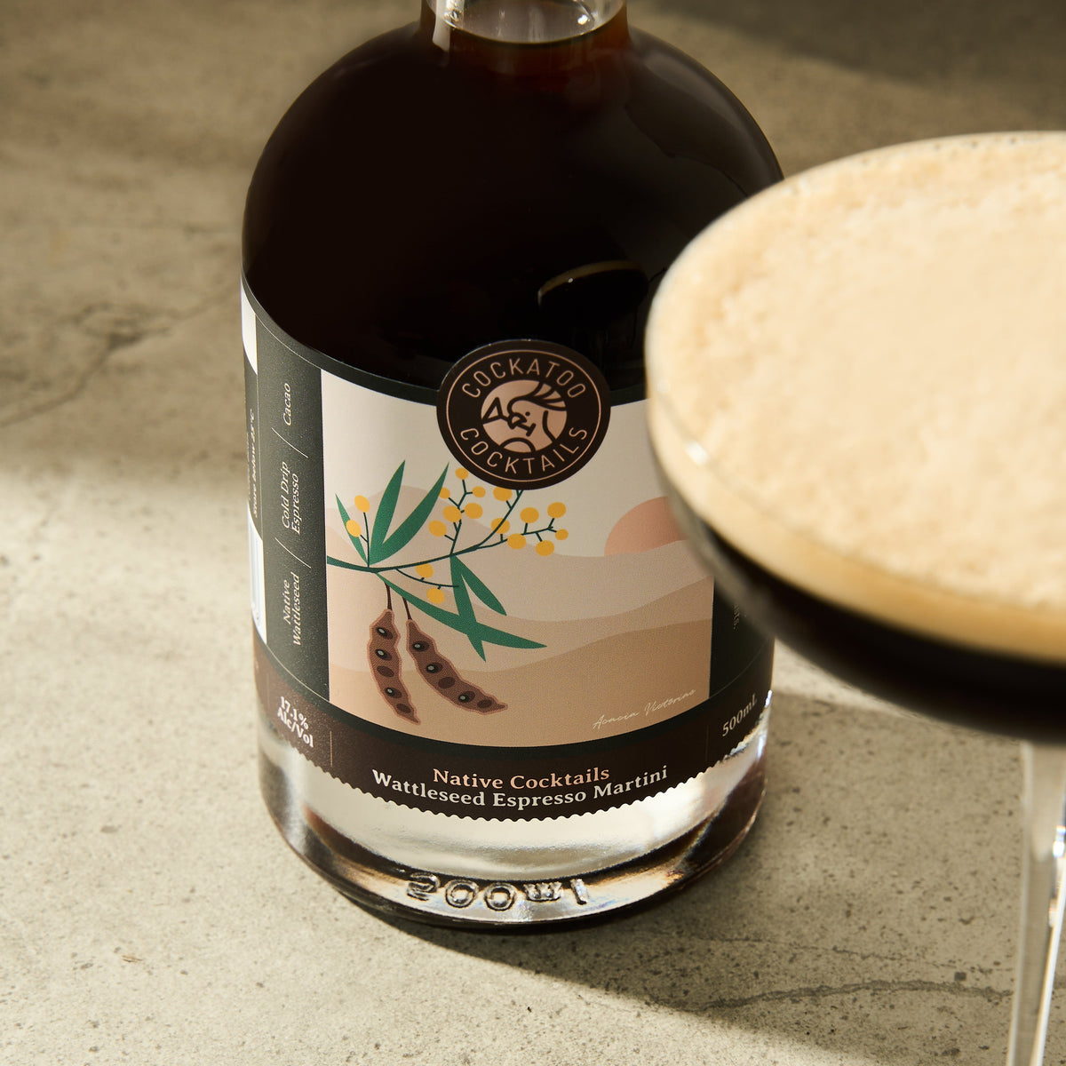 A bottle of Cocktails Cocktails Native Cocktails Wattleseed Espresso Martini next to a filled coupe glass with a thick cream foam head, set on a textured gray stone surface.
