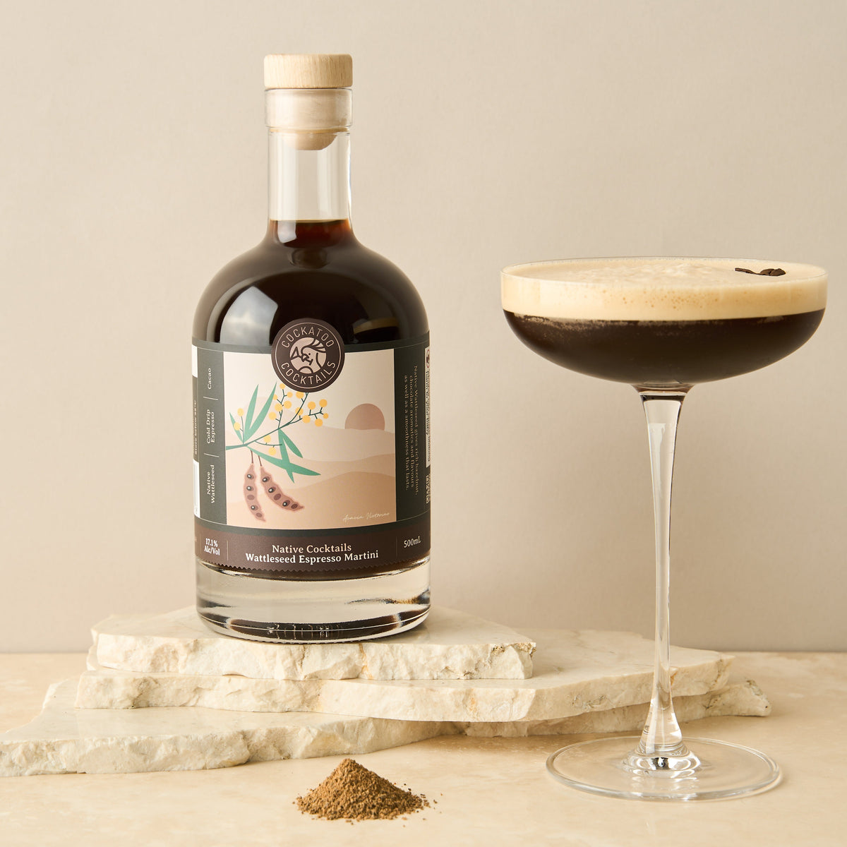 A bottle of Wattleseed Espresso Martini Cocktail placed next to a filled cocktail glass on a marble surface, with a small pile of wattleseed in front.